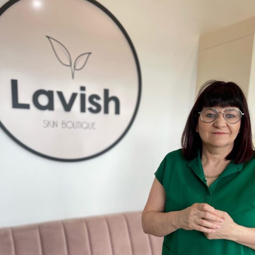Lavish sskin boutique robina dmk skin clinic new year beauty salon gold coast A mature female beautician wearing spectacles and dressed in black looks directly at the camera with the Lavish Skincare Boutique logo on the wall behind her.
